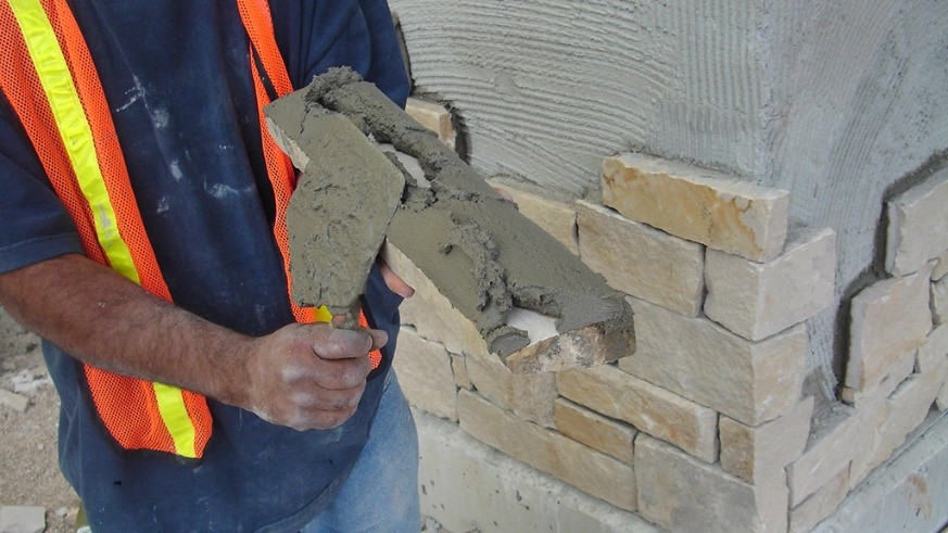 A skilled stonemason carefully installs a natural stone veneer around a fireplace, precisely fitting each unique piece of stone to create a beautiful, rustic surround. The craftsmanship highlights the textured variety of the stones in shades of gray, brown, and beige, enhancing the warmth and inviting ambiance of the room.