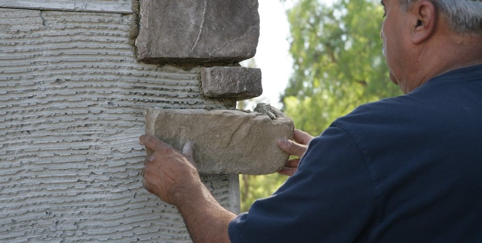 The thick, gray mixture is perfectly blended, showing a smooth, consistent texture ideal for bonding stone pieces to surfaces. In the background, a few stone veneer pieces and a trowel rest on a workbench, indicating the ongoing masonry work. This image emphasizes the essential role of high-quality premixed mortar in ensuring durable and secure installation of stone veneer projects.