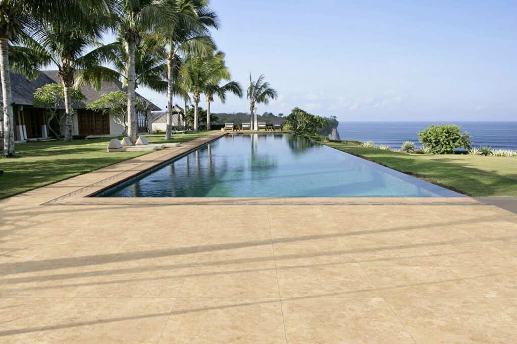 Luxurious Tuscany Beige travertine patio surrounding a long swimming pool, perfect for outdoor relaxation and entertainment, highlighting durable and stylish travertine pavers from Peninsula Building Materials.