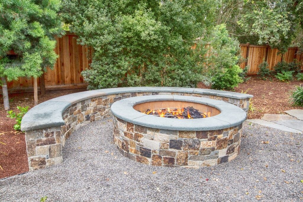 Round outdoor fire pit made of natural stone, featuring a curved natural stone bench, creating a perfect gathering spot for cozy evenings and outdoor entertainment.