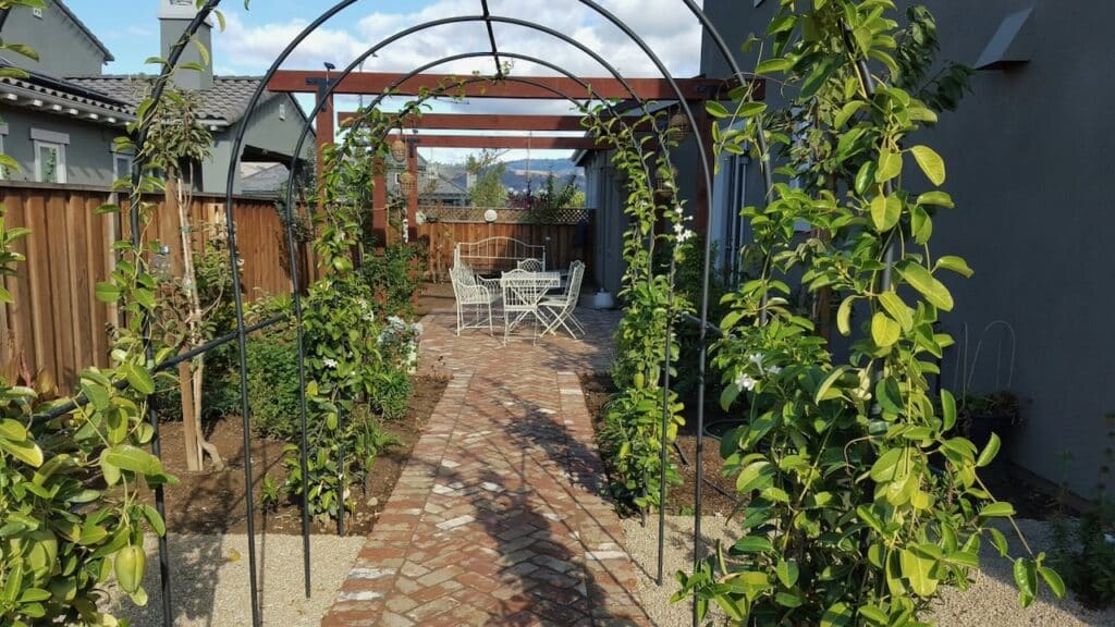 A charming garden walkway made of red and tan brick pavers in a herringbone pattern, bordered by lush greenery and climbing plants on metal arch trellises, leading to a cozy outdoor seating area with white wrought-iron furniture and a pergola.