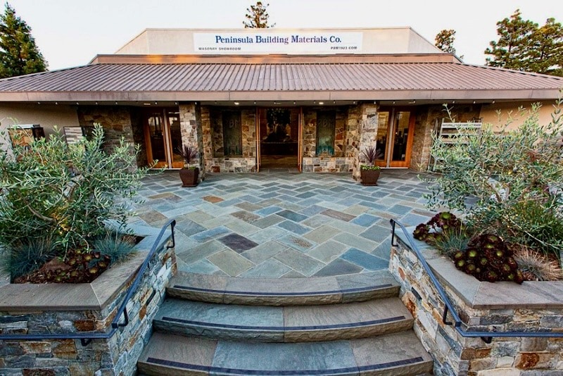 The entrance of Peninsula Building Materials showroom, featuring a spacious patio with elegant natural stone paving and lush landscaping.