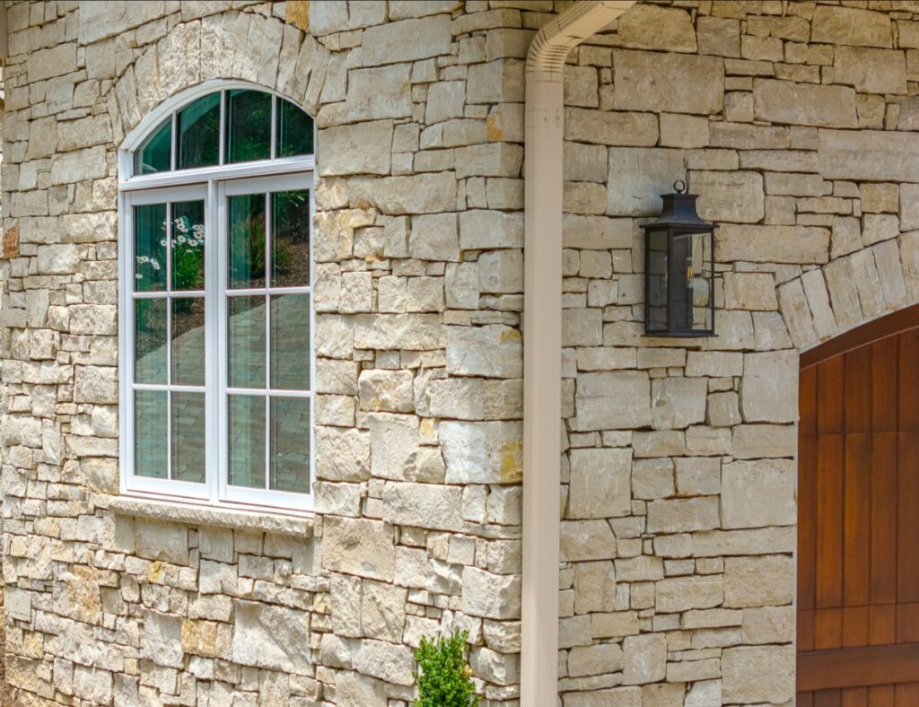 Rustic home facade showcasing Jute Cloth Country Squire stone veneer with natural buff hues and gray striations, complemented by arched windows and elegant lantern lighting.