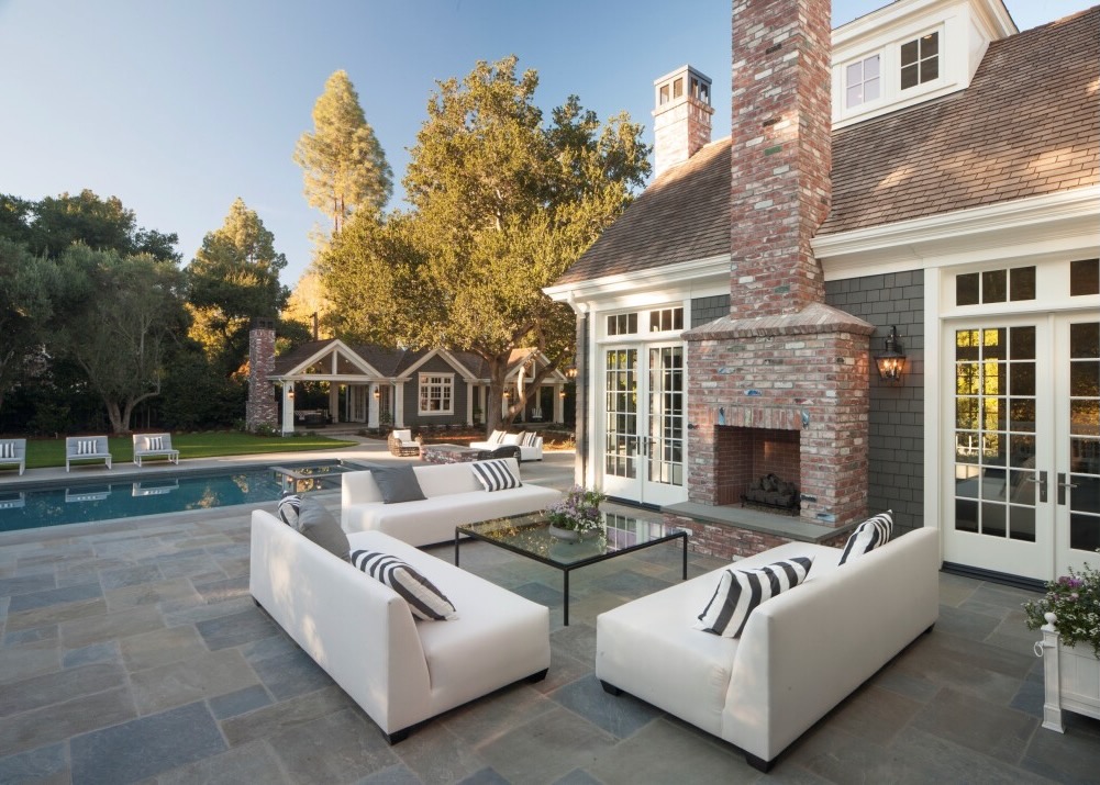 Classic outdoor brick fireplace on a spacious patio next to a pool, surrounded by elegant white outdoor sofas for relaxing by the fire.