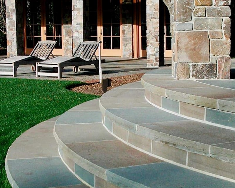 A luxurious outdoor patio with curved bluestone steps leading to a covered seating area, demonstrating the durability and fire resistance of natural stone.