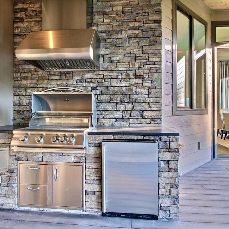 An outdoor kitchen featuring a built-in stainless steel grill set against a backdrop of stacked stone veneer, combining fire resistance with design sophistication.