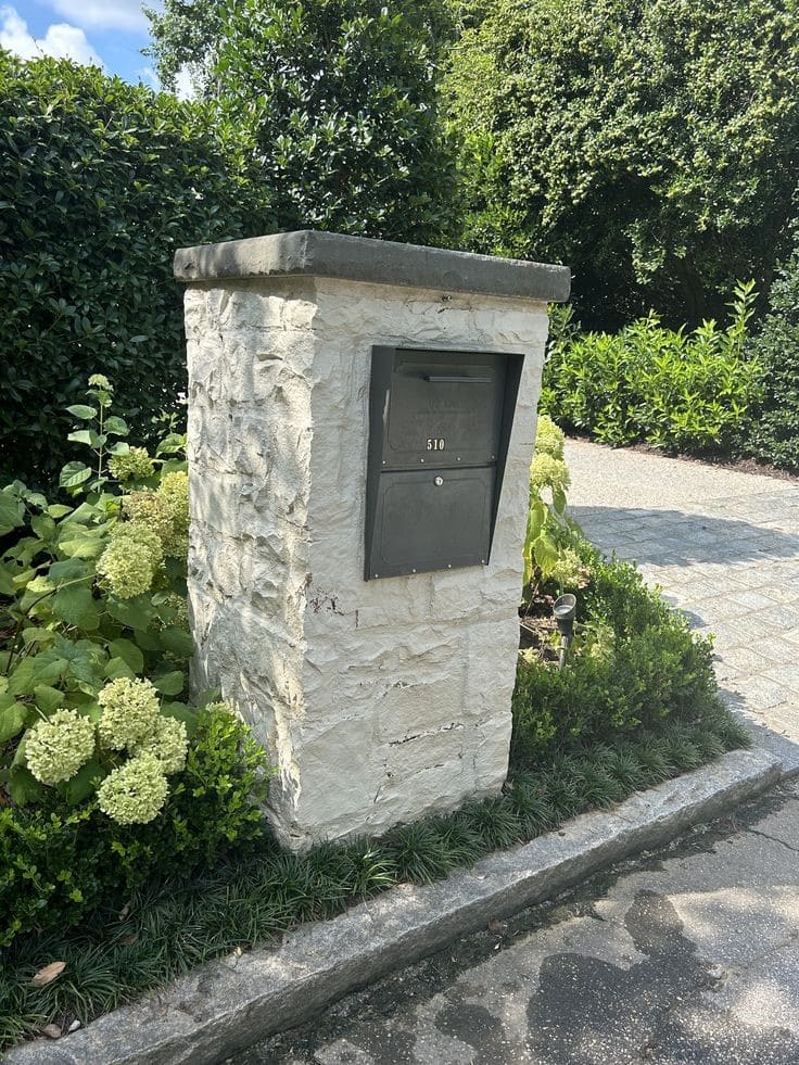 Light-colored stone mailbox post with black inset mailbox and dark stone cap, surrounded by hydrangeas and manicured greenery.