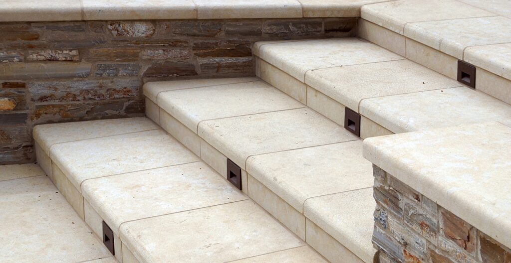 Outdoor staircase with cream-colored stone treads and risers, featuring rounded front edges and a matching stone veneer retaining wall.