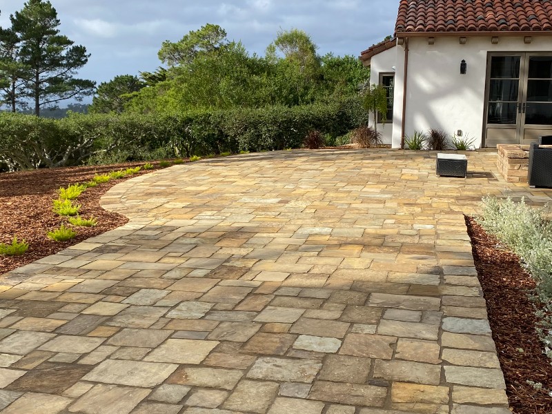 Expansive stone patio made of multi-colored rectangular pavers leading to a stucco home with a tile roof and surrounding landscaping.