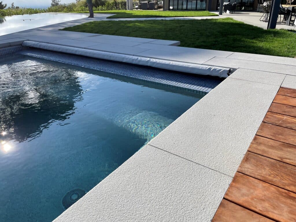 Modern swimming pool with light textured stone coping and surrounding patio next to a warm wood deck and lawn.