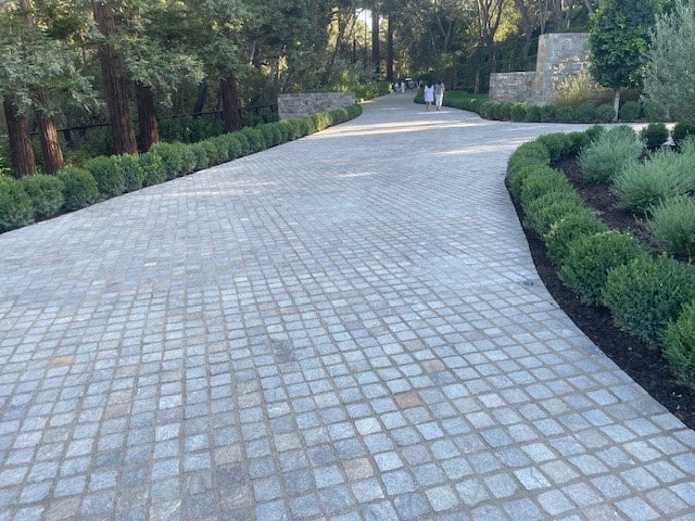 Long residential driveway paved with small square stone cobbles, curving through landscaped greenery and mature trees.